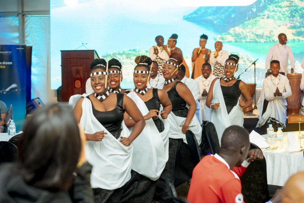 Female traditional dancers perform at an event in BK Arena. They are dressed in elegant Rwandan attire with flowing skirts and beaded accessories. They gracefully move in sync, showcasing the beauty of Rwandan cultural dance against the backdrop of a vibrant crowd and stage lights.