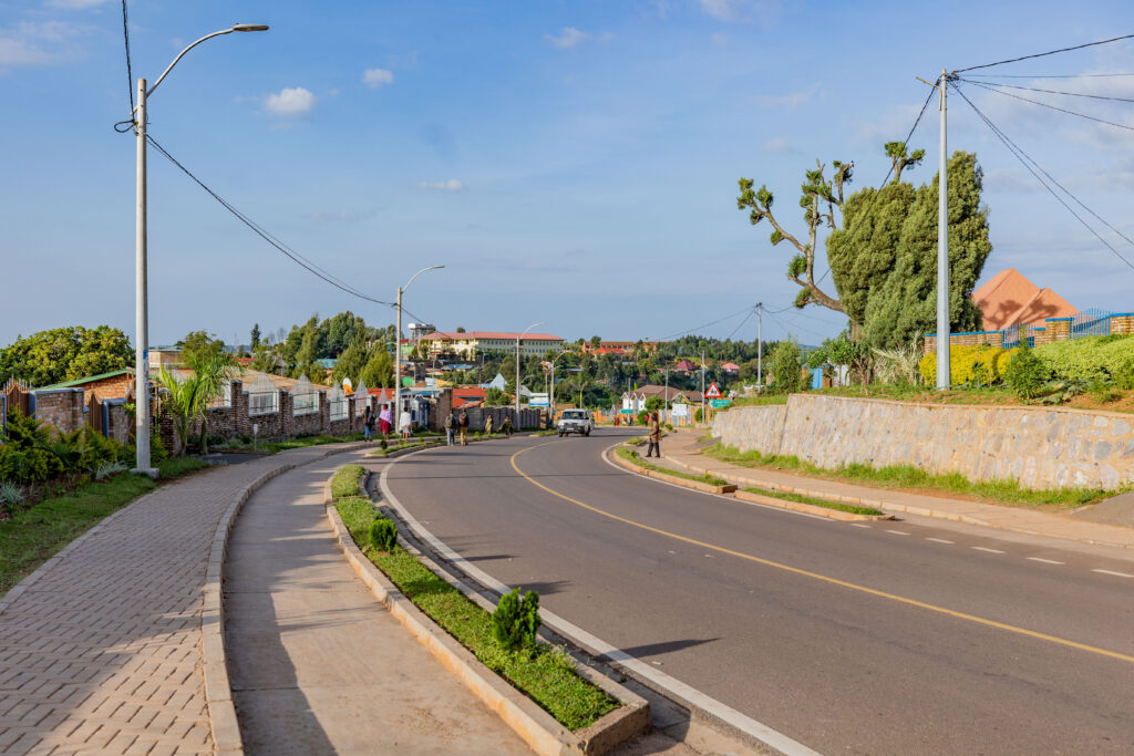  A quiet, well-maintained road in Rwanda, curving through a residential area with sidewalks, greenery, and houses in the background. A few pedestrians walk along the sidewalk.