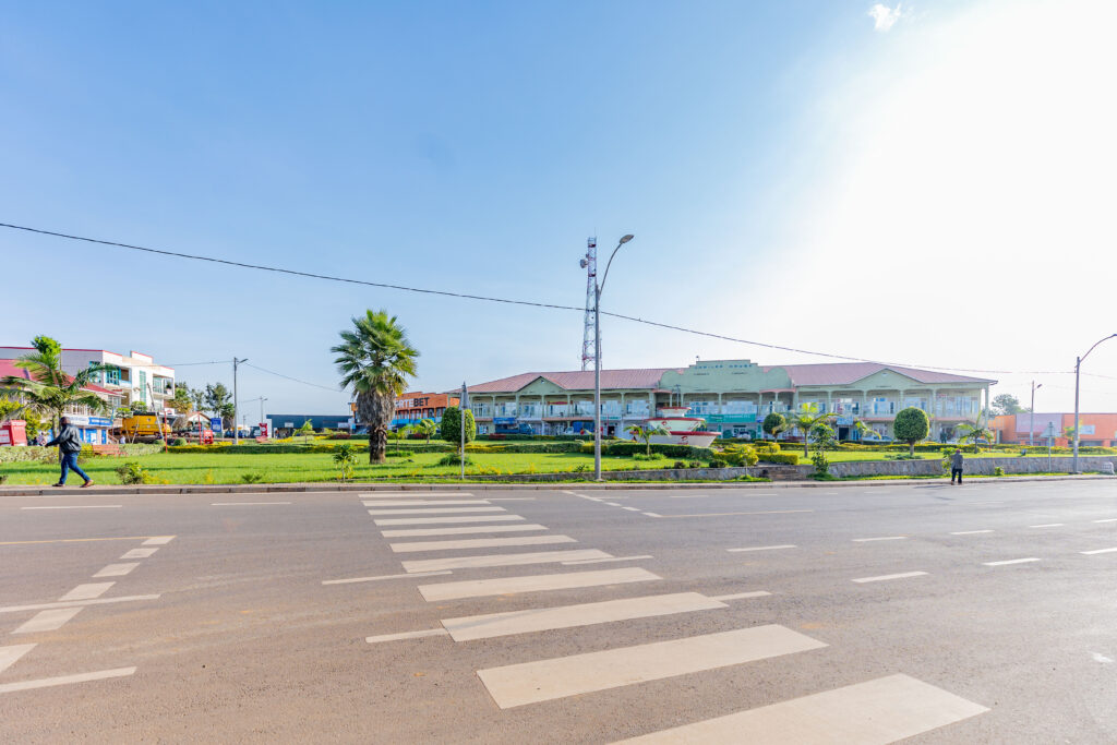 A wide road with a pedestrian crossing in front of a commercial area in Rwanda. The center of the road has a landscaped island with palm trees, and various shops and buildings are visible.