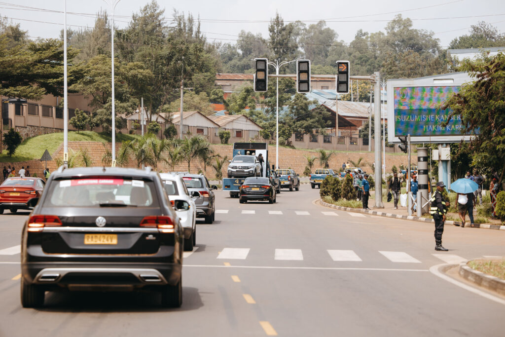A busy urban street in Rwanda with multiple cars, pedestrians, and a security officer. A digital billboard displays a message in the local language, and the road is lined with trees and buildings.