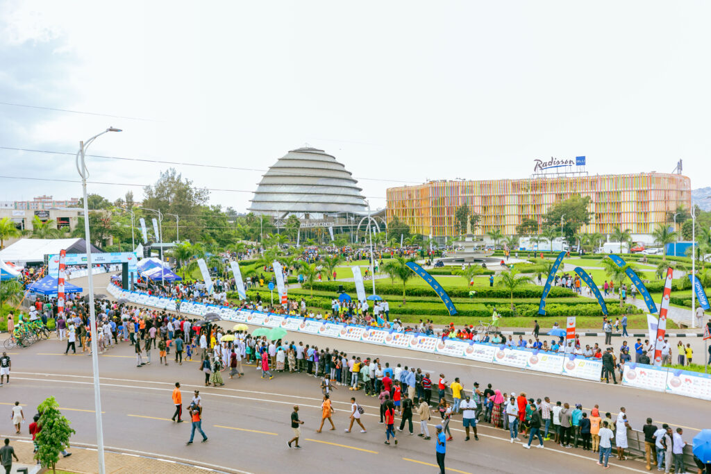 A large public event in Kigali, Rwanda, with a crowd gathered along a road near the iconic Kigali Convention Centre and Radisson Blu Hotel. The event features banners, tents, and spectators.