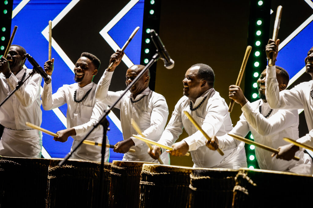 "Intore drummers performing at an event in BK Arena, dressed in traditional Rwandan attire with vibrant robes and headdresses, energetically beating drums in unison. The arena's bright lights and cheering crowd add to the lively atmosphere.